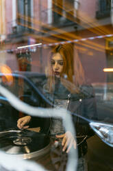 Durch das Fenster einer Frau, die einen alten Plattenspieler abspielt, während sie in einem Schallplattengeschäft in der Nähe einer Straße mit Auto steht - ADSF32773