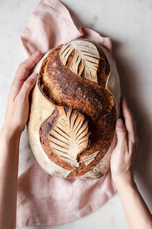 Top view of crop anonymous person with freshly baked bread with creative leaf pattern on white background in light kitchen - ADSF32776