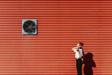 Woman wearing knit hat by red corrugated wall on sunny day - EGAF02467