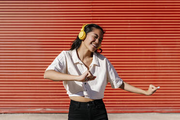 Happy woman with headphones dancing in front of wall - EGAF02475