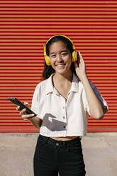 Frau mit Kopfhörern und Mobiltelefon vor roter Wand an sonnigem Tag - EGAF02478