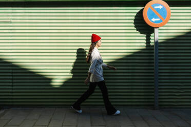 Young woman walking by corrugated wall on sunny day - EGAF02497