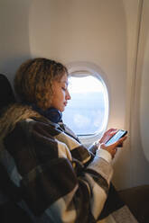 Frau benutzt Smartphone in der Nähe des Fensters im Flugzeug sitzend - MRAF00800