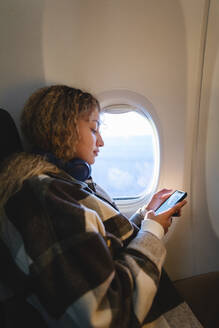 Woman using smart phone sitting near window in airplane - MRAF00800