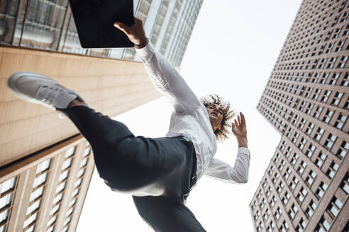 Excited businessman holding digital tablet jumping amidst office buildings - GUSF06710