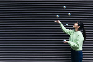 Young woman juggling globes by black corrugated wall - ASGF01903