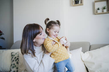 Smiling mother holding playful daughter on sofa - EBBF05090