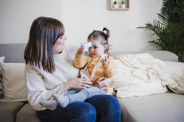 Lächelnde Mutter betrachtet ein süßes kleines Mädchen, das ein Infrarot-Thermometer auf dem Sofa hält - EBBF05093