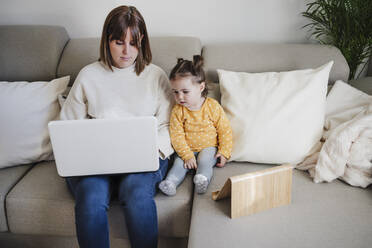 Mother using laptop by daughter with tablet PC on sofa - EBBF05099