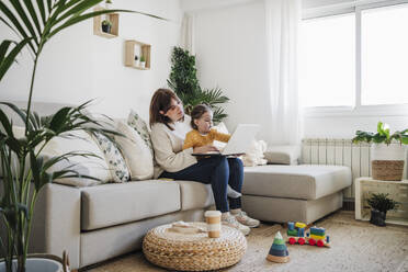 Mother sitting with daughter and using laptop in living room - EBBF05103