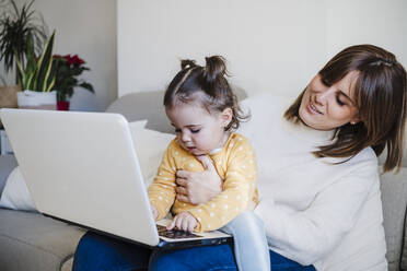 Lächelnde Mutter betrachtet ihre Tochter, die auf dem Sofa einen Laptop benutzt - EBBF05104