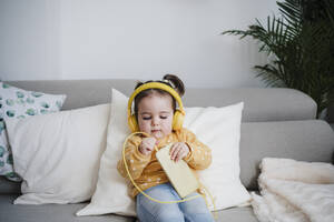 Baby girl putting headphones in mobile phone on sofa - EBBF05107