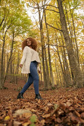 Smiling young woman walking on leaves in autumn forest - VEGF05229