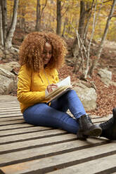 Young woman with curly hair reading book on bridge in autumn forest - VEGF05240