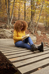 Smiling young woman reading book in autumn forest - VEGF05241