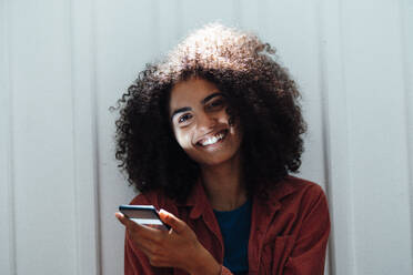Smiling woman with smart phone leaning on white wall - KNSF09234