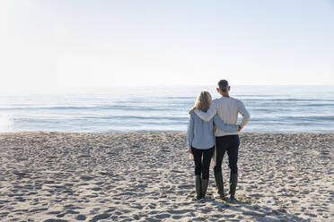 Mann mit Arm um Frau, die auf das Meer blickt, steht auf Sand - EIF02557