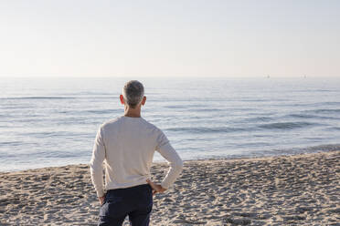 Mann mit Hand auf der Hüfte am Strand stehend und aufs Meer blickend - EIF02578