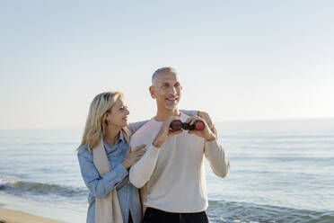 Lächelnde Frau mit Arm um ihren Mann, der ein Fernglas am Strand hält - EIF02587