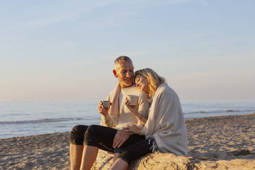 Paar mit Tee sitzen auf Baumstamm am Strand bei Sonnenuntergang - EIF02620