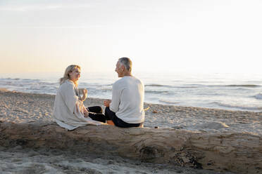Paar im Gespräch miteinander auf einem Baumstamm am Strand sitzend - EIF02623