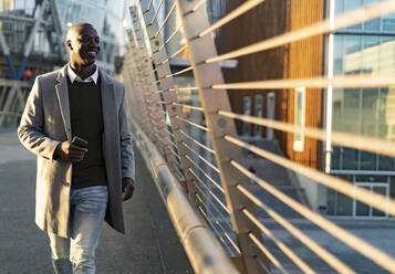 Lächelnder Geschäftsmann mit Smartphone, der in der Nähe des Geländers einer Brücke spazieren geht - GIOF14383