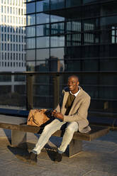 Lächelnder Geschäftsmann mit Smartphone sitzt auf einer Bank vor einem Gebäude - GIOF14386