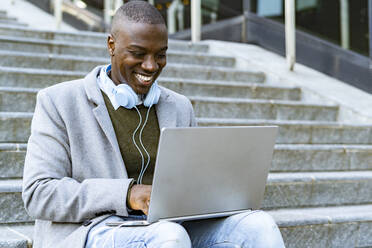 Smiling businessman with headphones using laptop on steps - GIOF14420