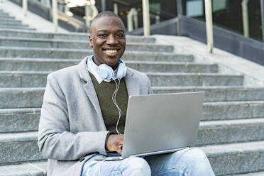 Smiling businessman with headphones and laptop sitting on steps - GIOF14421