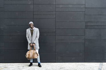 Smiling businessman with duffel bag standing in front of black wall - GIOF14423