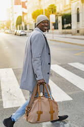 Smiling businessman with duffel bag crossing road in city - GIOF14424