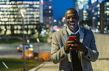 Geschäftsmann mit Smartphone lachend in der Stadt bei Nacht - GIOF14464
