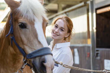 Smiling woman looking at horse in stable - EIF02638