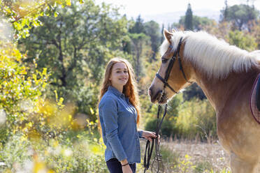 Smiling woman standing with horse at meadow on sunny day - EIF02656