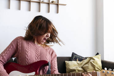 Young woman screaming and playing guitar on sofa - ASGF01921