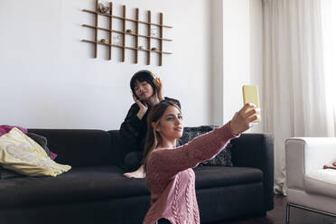 Woman taking selfie through smart phone with friend listening music on sofa - ASGF01927