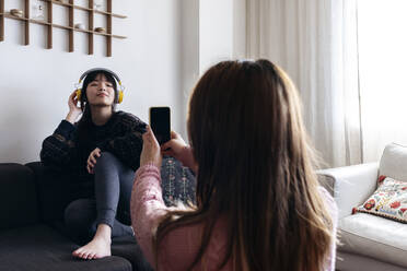 Woman photographing friend through mobile phone at home - ASGF01928