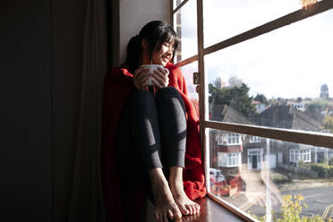 Smiling young woman holding coffee mug sitting on sill looking out from window at home - ASGF01945