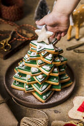 Hand eines Jungen an einem Weihnachtsbaum, der aus gestapelten Keksen in einem Teller auf dem Tisch besteht - FLMF00739