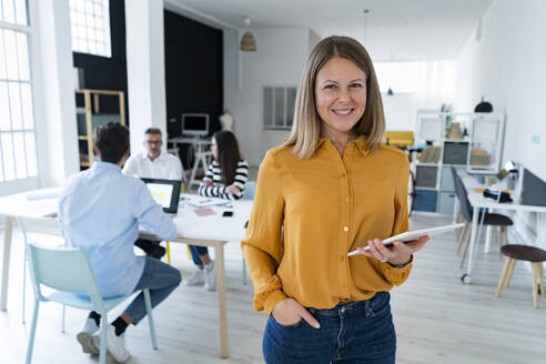 Businesswoman with hand in pocket holding tablet PC with colleagues sitting at office - GIOF14525