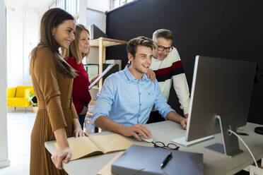 Junger Geschäftsmann an einem Desktop-PC mit Kollegen, die am Schreibtisch im Büro stehen - GIOF14542