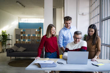 Businessman amidst colleagues looking at laptop at desk in office - GIOF14549