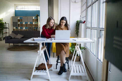 Businesswoman leaning by colleague using laptop at desk in office - GIOF14551