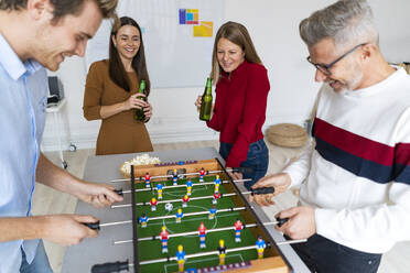 Smiling businesswomen with beer bottles looking at businessmen playing foosball in office - GIOF14586