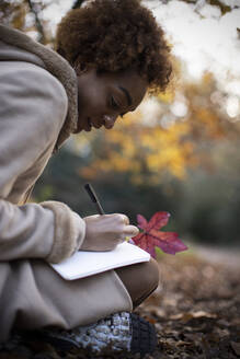 Young woman with autumn leaf writing in journal in park - CAIF31826