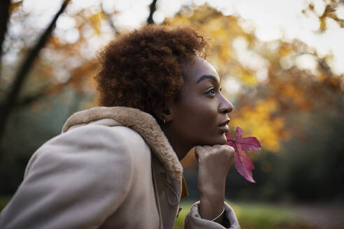 Thoughtful young woman with red autumn leaf in park - CAIF31850