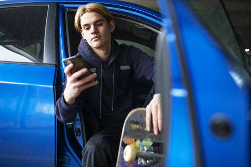 Jugendlicher mit Skateboard und Smartphone in der Autotür - CAIF31869