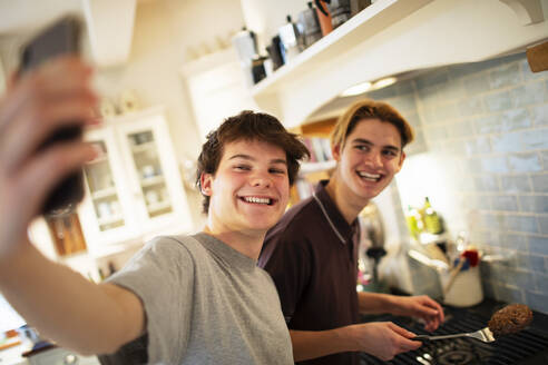 Happy teenage boys taking selfie and cooking in kitchen - CAIF31883
