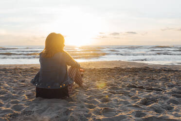 Back lit woman enjoying sunset sitting on sand looking at horizon - EGHF00286