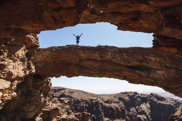 Young man with arms outstretched standing on natural rock arch, Gran Canaria, Spain - RSGF00749